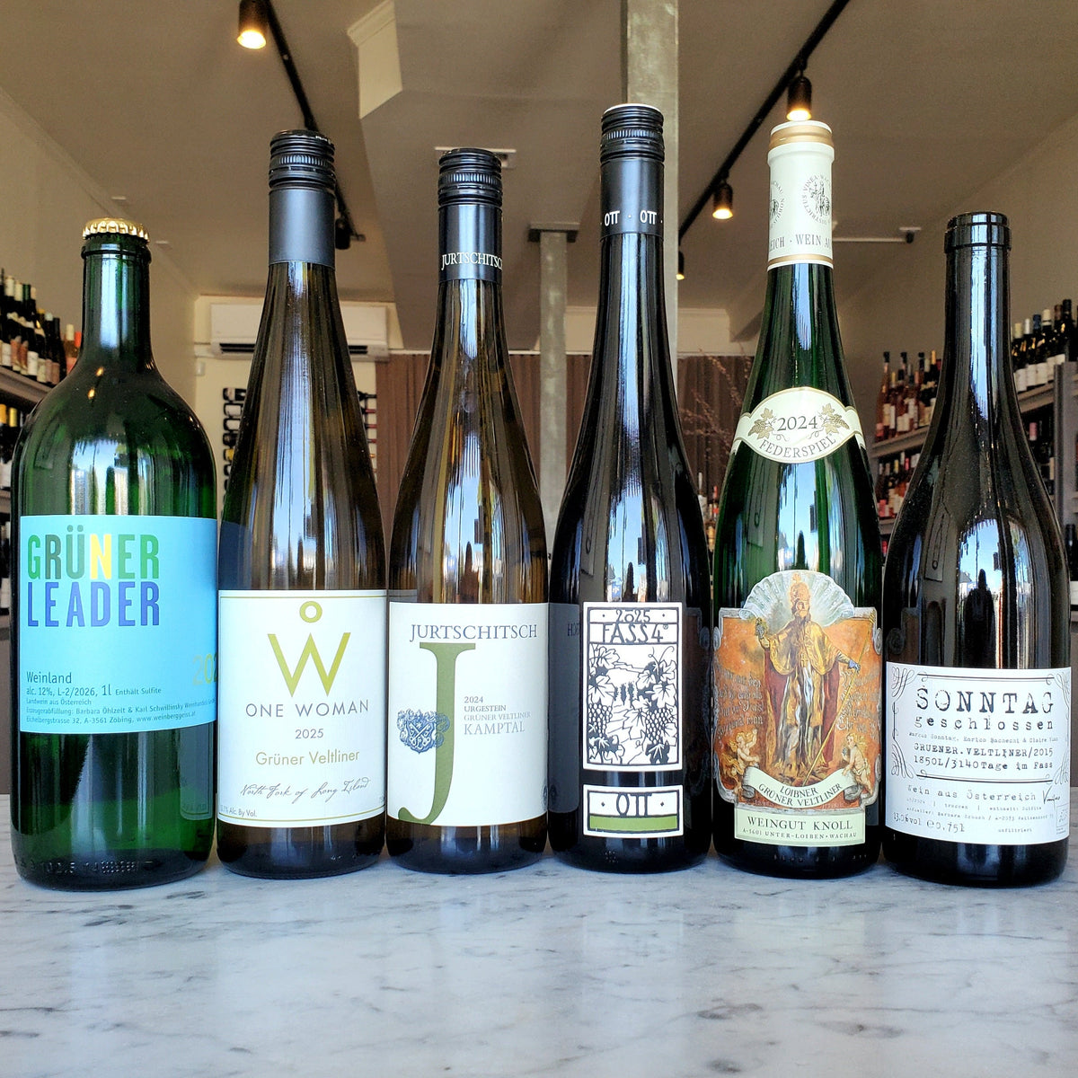 Six bottles of Austrian Gruner Veltliner wine lined up on a white marble countertop in a wine shop. The bottles include green and dark glass with colorful labels in blue, gold, black, and orange.