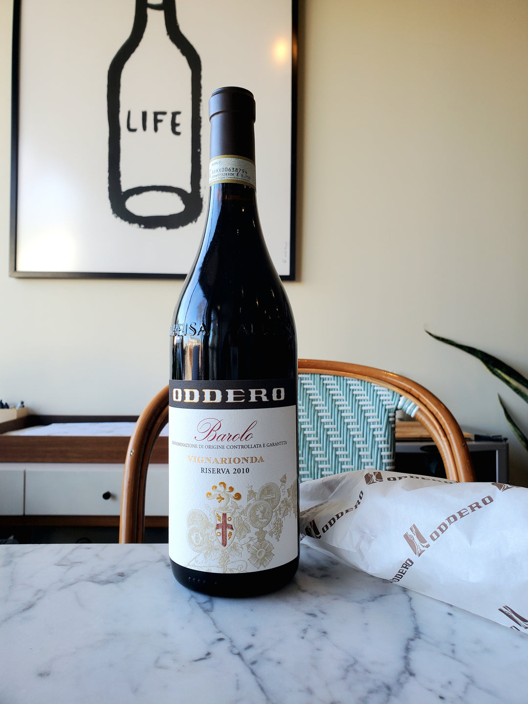 A bottle of Oddero Vignarionda Barolo, Italy 2010 red wine placed on a marble surface with a framed artwork 'LIFE' on the wall in the background.