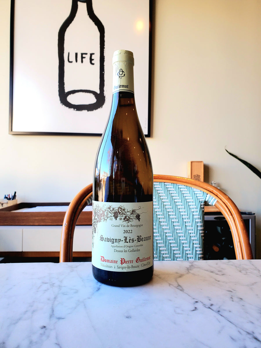 A bottle of Pierre Guillemot 'Dessus les Gollardes' Savigny-les-Beaune Blanc wine positioned on a marble surface with a wine glass in the background, set against a wall art with the phrase 'life'.